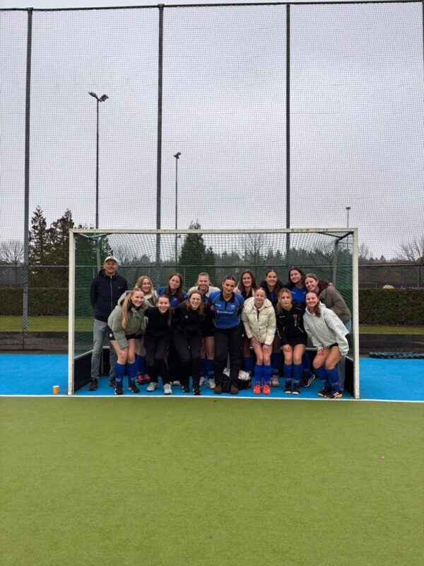 Groepsfoto van een hockeyteam op het veld bij het doel.